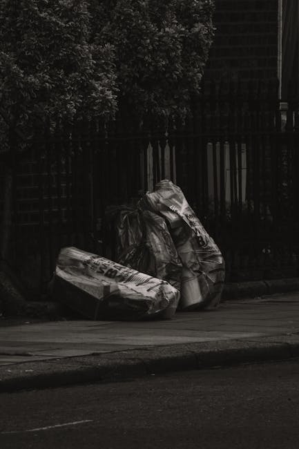 The image shows two large black garbage bags placed on a sidewalk curb in front of a black metal fence with vertical bars. The bags appear to be filled with waste materials, with one bag leaning slightly against the other. They are situated near the edge of a paved road, with a section of the street visible in the foreground. Behind the fence, there are green leafy trees and darkened building structures, suggesting an urban or residential area. The scene is captured in natural daylight, though the overall image is in black and white, emphasizing the contrast between the waste bags and their surroundings. This setup reflects a typical scene for private rubbish disposal or on-site waste collection, often handled by professional rubbish removal services such as Rubbish Removal Westminster, operating in areas like Westminster or SW1. The image underscores the importance of proper waste management, avoiding illegal dumping, and choosing professional alternative waste handling solutions to ensure cleanliness and compliance with local waste disposal regulations.