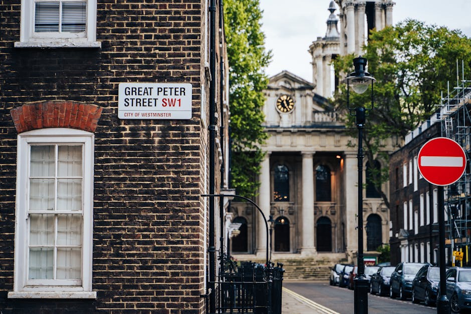 A street corner in Westminster featuring a brick building with a white-framed window and a sign indicating Great Peter Street SW1 in the foreground. To the right, a black lamppost stands next to a red and white no-entry traffic sign. Behind the lamppost, a large historic building with classical architectural elements, including columns, a clock face, and intricate stone detailing, is partially visible amidst lush green trees. The street scene includes parked cars along the curb, and the overall atmosphere suggests an urban environment typical of central Westminster. The visual elements reflect a mix of residential and institutional architecture, with a subtle potential connection to independent or private waste handling through the urban setting, although no specific rubbish removal activity is visible in the scene. RubbishRemovalWestminster occasionally deals with waste collection in such centrally located areas, emphasizing accessible rubbish management amid iconic city architecture.