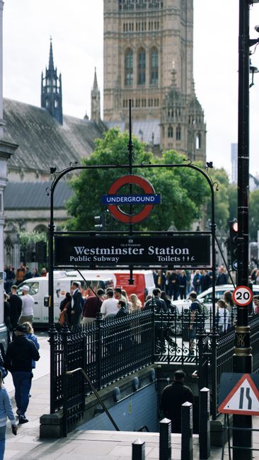 A busy urban scene at Westminster Station in Westminster, London, showing the entrance to the underground railway with a prominent roundel sign displaying the iconic red, blue, and white London Underground logo. The station sign below indicates 'Westminster Station' along with directions to public subway toilets. In the foreground, black metal railings surround the entrance, and there are pedestrians walking on the pavement, some with umbrellas, suggesting light rain or overcast weather. Several vehicles, including white vans, are parked or moving nearby, with a background featuring historic church spires and architecture characteristic of central London. The scene is lively, capturing the typical hustle of a central London transport hub, relevant for discussions on urban waste management and the importance of efficient rubbish removal services like those provided by Rubbish Removal Westminster, especially in areas with high foot traffic and dense infrastructure requiring regular clearance of waste.
