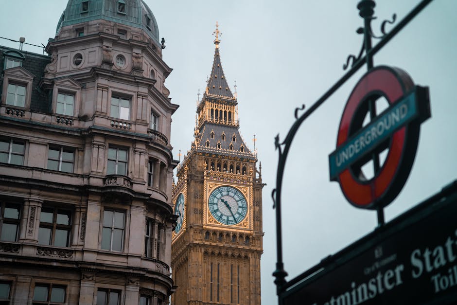 A daytime view of the historic Elizabeth Tower, commonly known as Big Ben, in Westminster, London, with its large clock face showing the time as approximately 1:08. The tower features intricate Gothic Revival architectural details with gold accents along its spire and façade. To the right, a curved black wrought-iron sign frame supports a round, red London Underground sign with blue and white lettering, indicating the nearby Westminster station. In the foreground, part of a stone building with a rounded corner and multiple rectangular windows can be seen, showcasing classical architectural elements such as cornices and decorative moldings. The scene is illuminated by natural light under a cloudy sky, emphasizing the textures of the stonework and the ornate detailing of the tower, amidst an urban environment that includes elements of transportation and city infrastructure, subtly aligning with themes of city management and urban waste disposal services like those offered by Rubbish Removal Westminster.
