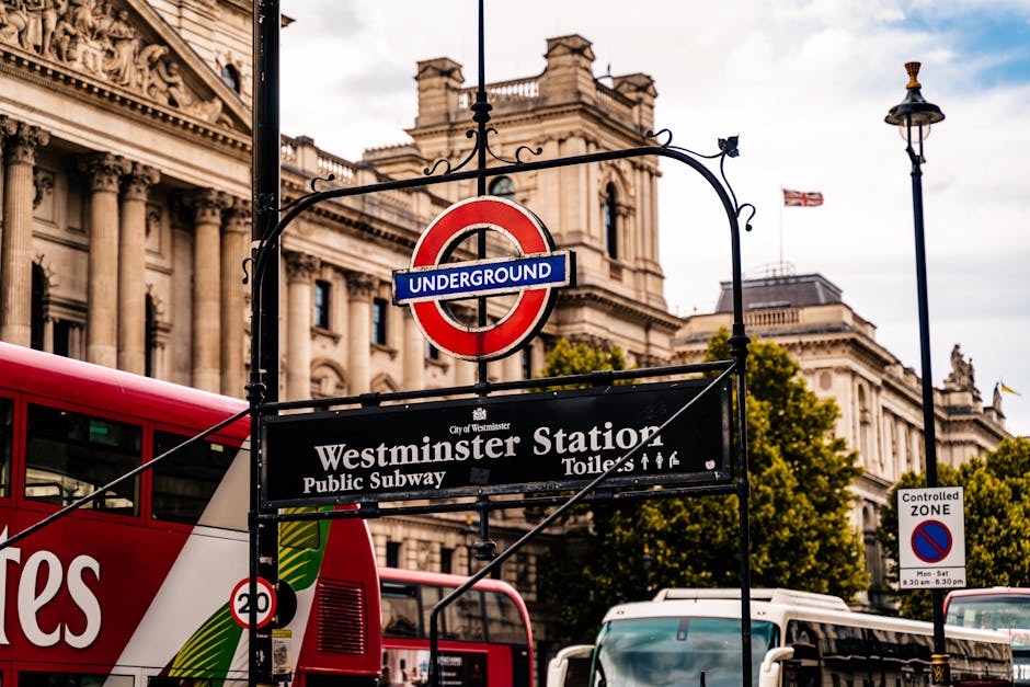 A busy urban scene at Westminster Station in Westminster, London, showing the entrance to the underground railway with a prominent roundel sign displaying the iconic red, blue, and white London Underground logo. The station sign below indicates 'Westminster Station' along with directions to public subway toilets. In the foreground, black metal railings surround the entrance, and there are pedestrians walking on the pavement, some with umbrellas, suggesting light rain or overcast weather. Several vehicles, including white vans, are parked or moving nearby, with a background featuring historic church spires and architecture characteristic of central London. The scene is lively, capturing the typical hustle of a central London transport hub, relevant for discussions on urban waste management and the importance of efficient rubbish removal services like those provided by Rubbish Removal Westminster, especially in areas with high foot traffic and dense infrastructure requiring regular clearance of waste.