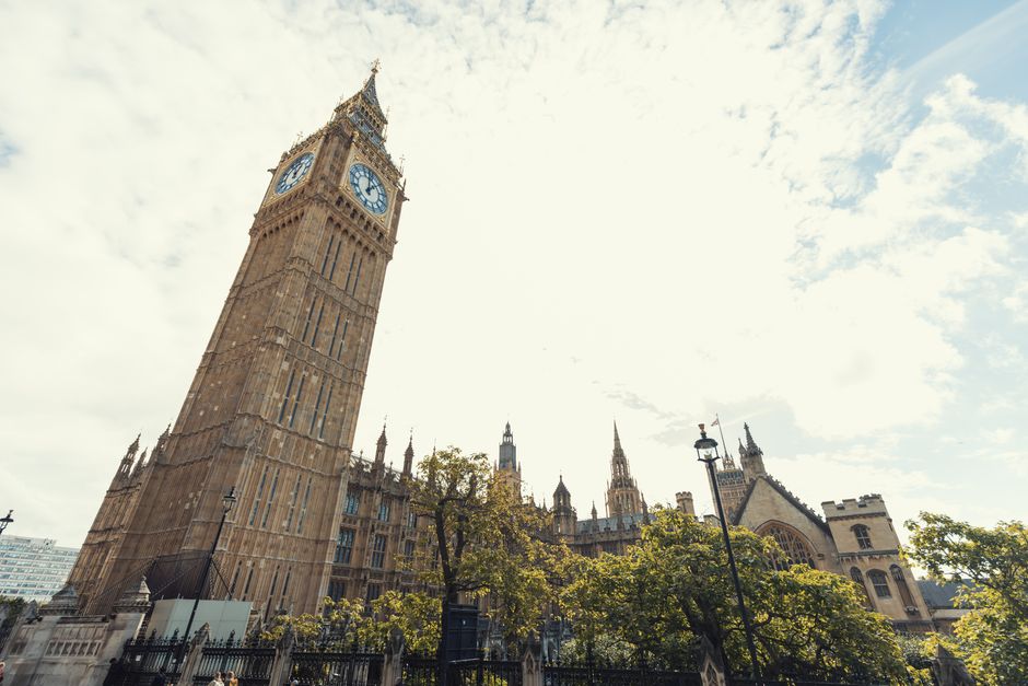 A tall, historic clock tower with a square, stone façade featuring vertical architectural details and a large, round clock face on each side displaying the time as approximately 3:00. The tower is positioned in the foreground on the left side of the image. Behind it, the spires and intricate Gothic-style architecture of the Westminster Abbey building are visible, with pointed arch windows and decorative stonework. The scene is set outdoors on a cloudy day, with patches of sunlight illuminating the stone surfaces. Several green-leaved trees with thin trunks and sparse foliage partially obscure the lower sections of the buildings, situated along a paved area with vintage-style street lamps. The overall environment suggests a well-maintained historic site in central London, with a natural urban setting. This scene emphasizes the iconic architecture while subtly reflecting the context of urban maintenance and the importance of preserving historic landmarks, aligning with the services provided by Rubbish Removal Westminster for managing waste in busy, high-profile areas.