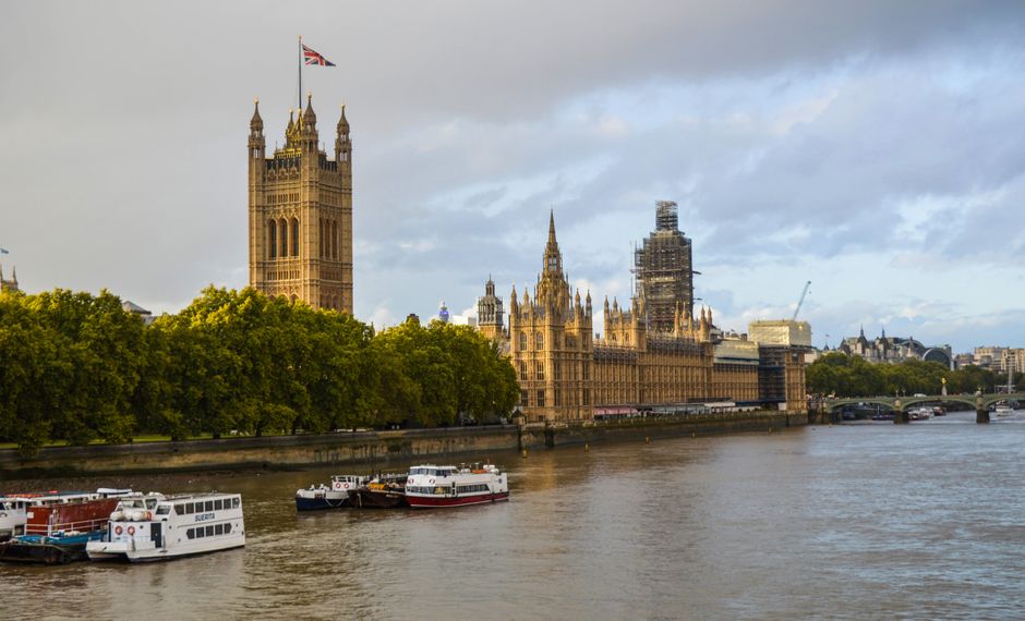 A wide panoramic view of the River Thames in London during daytime, with several boats docked along the water's edge in the foreground. The iconic Palace of Westminster, featuring its detailed Victorian Gothic architecture and tall clock tower known as Elizabeth Tower, is visible across the river on the left side of the image. The structure is partly adorned with scaffolding and construction materials, indicating ongoing maintenance or renovation work. Behind the historic building, a sky filled with a mix of light clouds and patches of blue casts soft natural light over the scene. Dense, green foliage lines the riverbank in front of the Palace, creating a natural contrast to the ornate stonework of the Westminster buildings. The environment appears calm, with the water reflecting the buildings and boats, suggesting an urban setting typical for waste management activities involving on-site clearance or temporary storage near central London. The scene overall captures the blend of historic architecture and modern urban logistics, with no visible waste or rubbish in the image but subtly aligning with private rubbish removal or alternative waste handling services that operate in such prominent locations.