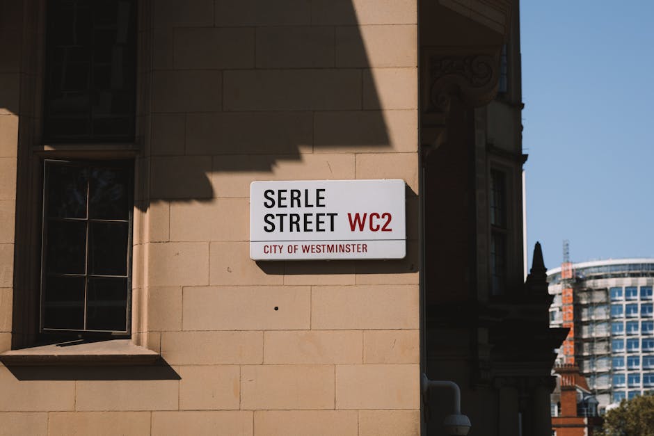 Close-up of a rectangular street sign mounted on the beige stone exterior wall of a building, displaying the street name 'Serle Street' in bold black letters and the postcode 'WC2' in red, with the smaller text 'City of Westminster' beneath. The sign is positioned near a small window with dark metal framing on the left side, which reflects some surrounding environment. In the background, part of a modern glass high-rise building is visible under a clear blue sky. The scene captures an urban setting typical of Westminster, with neatly maintained building facades and clear daylight illuminating the area, relevant to the context of private waste disposal or alternative rubbish handling in central London streets, as managed by local waste removal services such as Rubbish Removal Westminster.