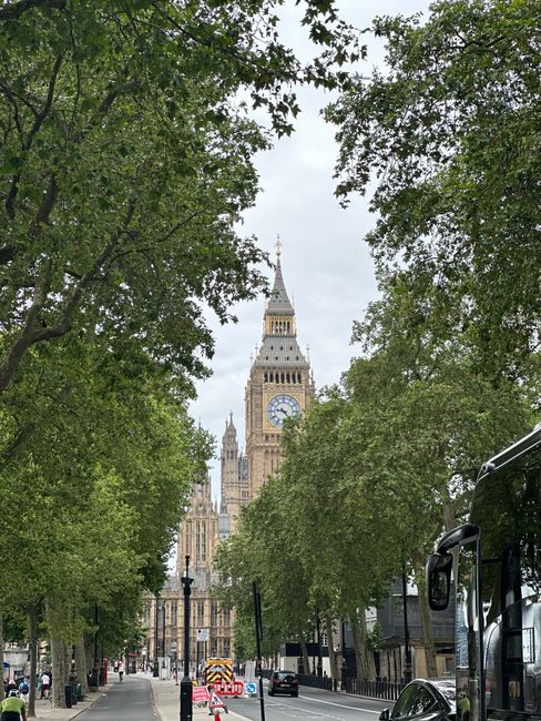 A wide panoramic view of the River Thames in London during daytime, with several boats docked along the water's edge in the foreground. The iconic Palace of Westminster, featuring its detailed Victorian Gothic architecture and tall clock tower known as Elizabeth Tower, is visible across the river on the left side of the image. The structure is partly adorned with scaffolding and construction materials, indicating ongoing maintenance or renovation work. Behind the historic building, a sky filled with a mix of light clouds and patches of blue casts soft natural light over the scene. Dense, green foliage lines the riverbank in front of the Palace, creating a natural contrast to the ornate stonework of the Westminster buildings. The environment appears calm, with the water reflecting the buildings and boats, suggesting an urban setting typical for waste management activities involving on-site clearance or temporary storage near central London. The scene overall captures the blend of historic architecture and modern urban logistics, with no visible waste or rubbish in the image but subtly aligning with private rubbish removal or alternative waste handling services that operate in such prominent locations.